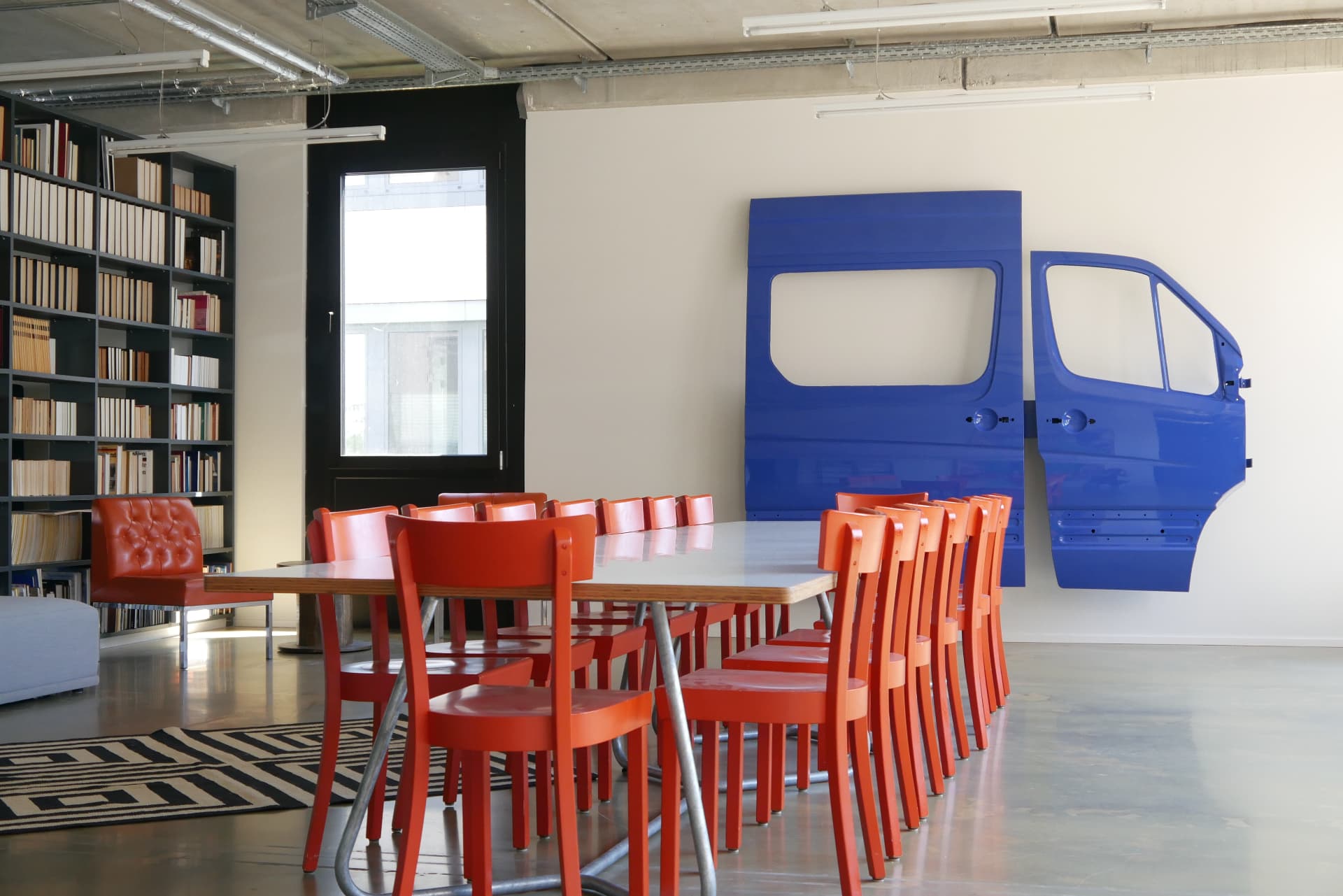 Library with book wall, long table, red chairs and a blue van-door artwork